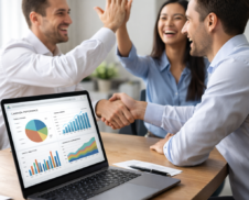 Professional team celebrating successful campaign results with handshake and high-five while analytics charts are visible on a laptop in a modern office
