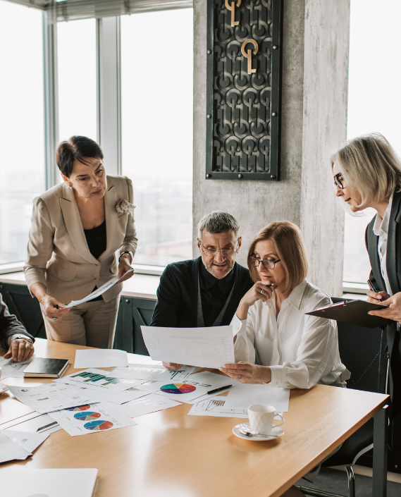 A group of four diverse professional colleagues reviewing business charts, pie graphs, and data reports on a wooden conference table in a modern office.